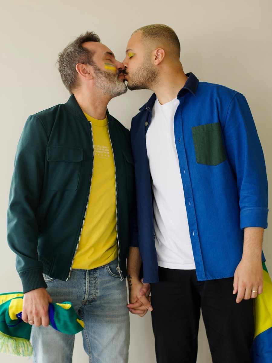 Imagem com fundo bege. No centro, dois homens de pé usando look para assistir aos jogos da Copa 2022. Eles estão se beijando.