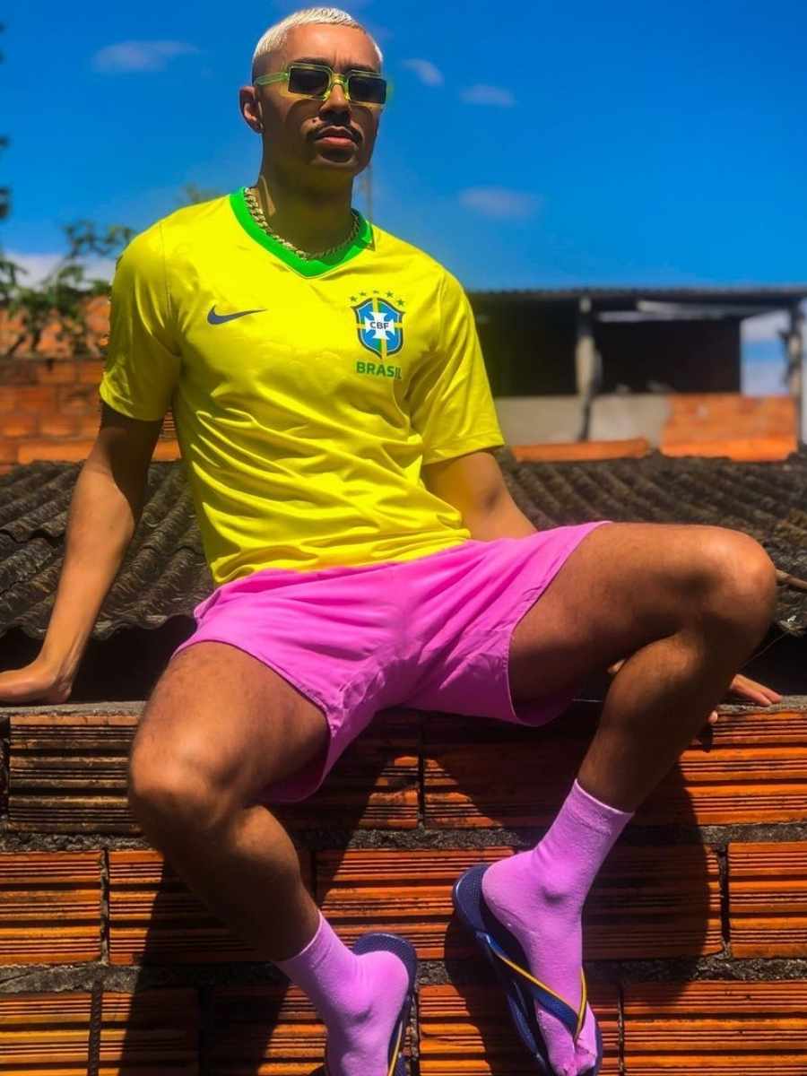 Imagem de homem sentado em muro de tijolo, usando camiseta amarela do Brasil com short rosa, meia rosa e sandália.
