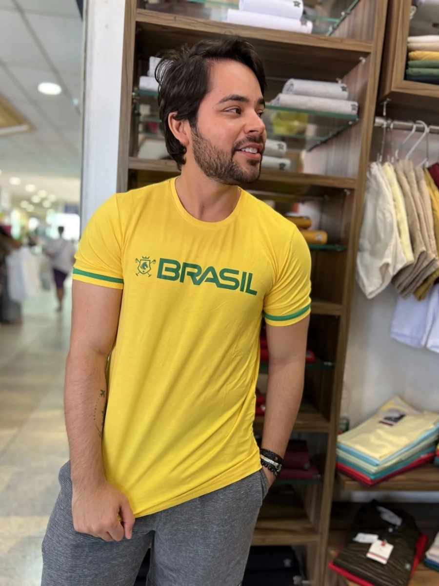Imagem de homem encostado em prateleira de loja, usando camiseta branca com estampa amarelo e verde, escrito "Brasil" no centro - looks masculinos para a Copa do Mundo.
