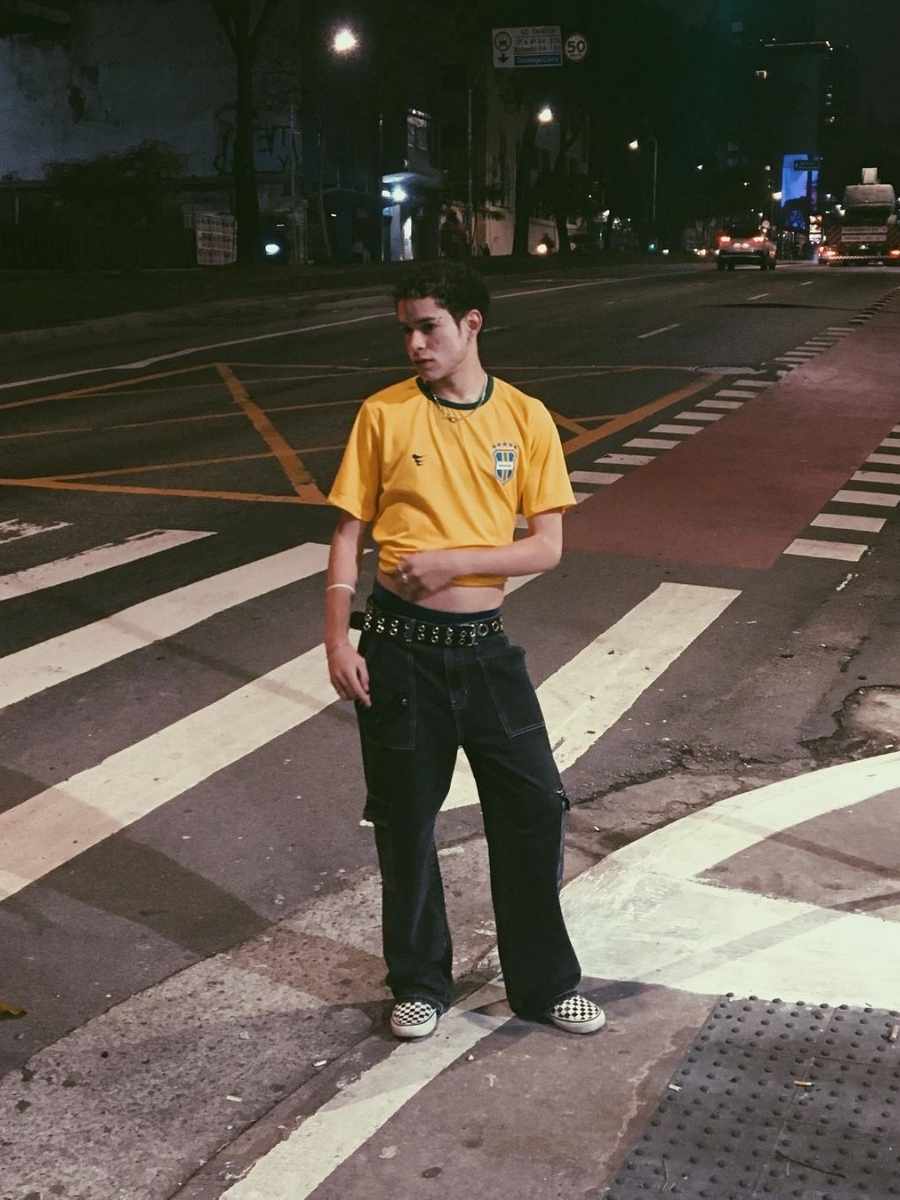 Imagem de homem de pé em uma rua, usando camiseta do Brasil + calça preta + cinto + sapatenis xadrez - looks masculinos para a Copa do Mundo.