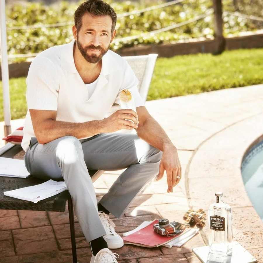 Imagem de ator Ryan Reynolds sentado em banco tomando sua bebida, gin Aviation American - famosos que ganham dinheiro vendendo bebida.
