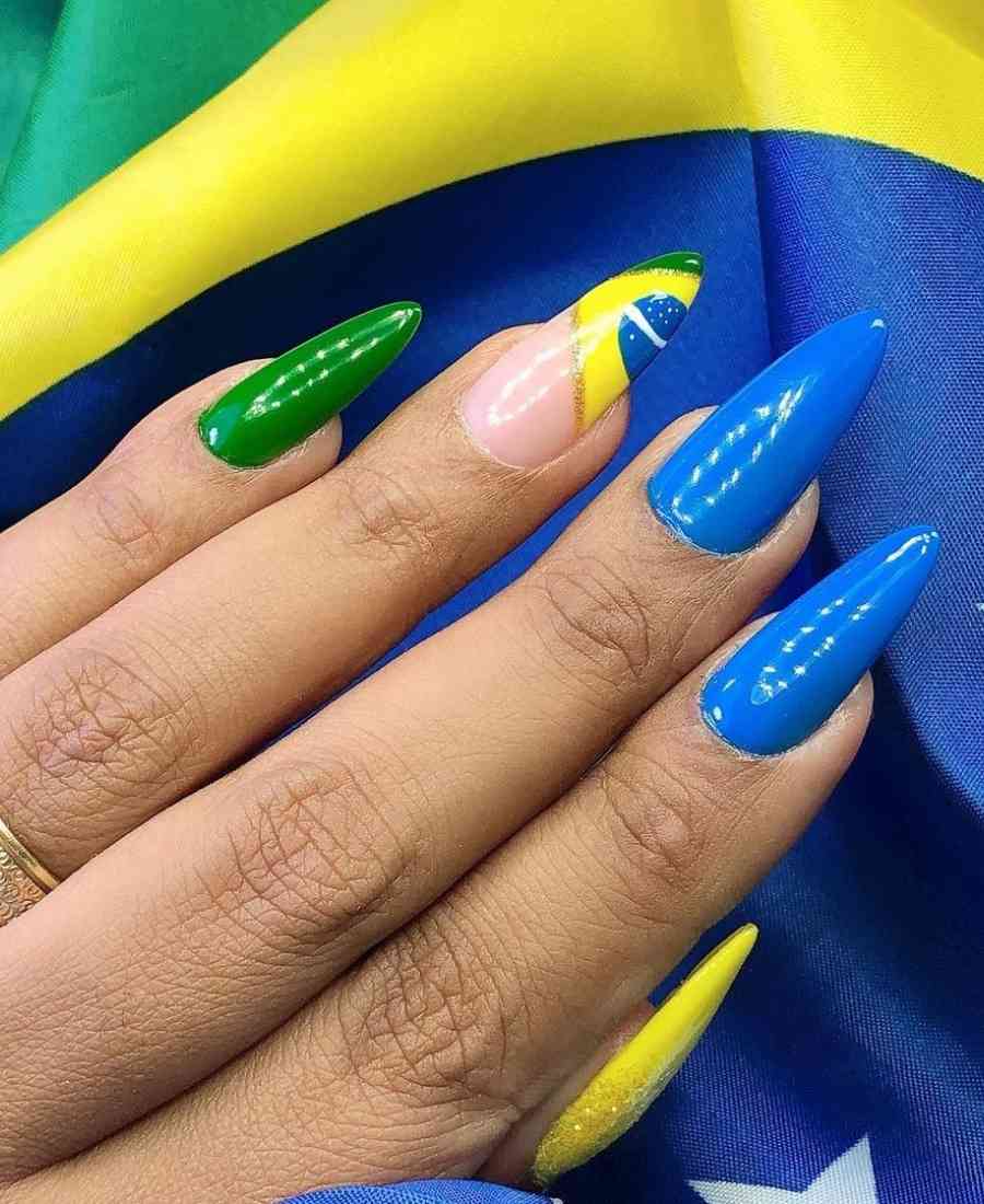 Imagem de mão com unhas decoradas para a Copa do Mundo 2022.  Cores usadas, verde, amarelo, azul e branco. Polegar, amarelo. Indicador e dedo médio, azul. Anelar, desenho da bandeira do Brasil. Mínimo, verde.