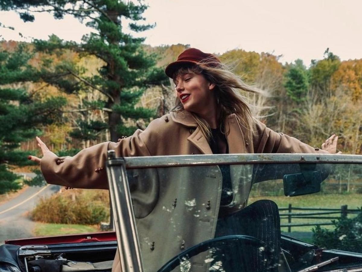 Imagem de Taylor Swift em carro luxuoso de capô aberto de pé com os braços abertos.