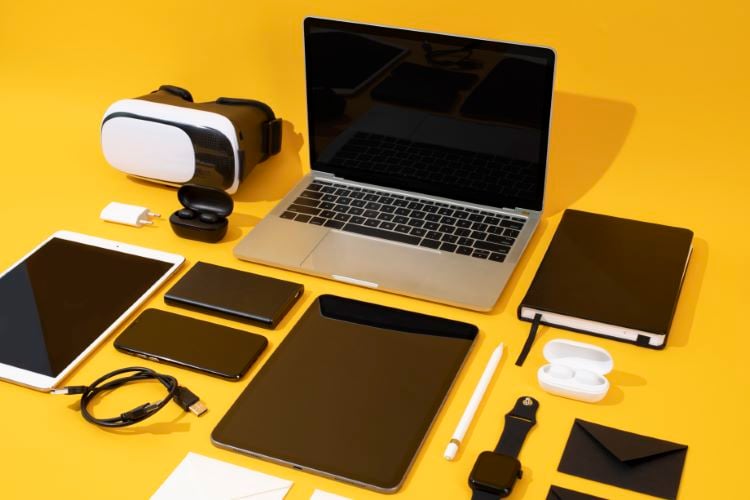 Mesa amarela com notebook, tablet, óculos de realidade virtual, celulares e relógio. 