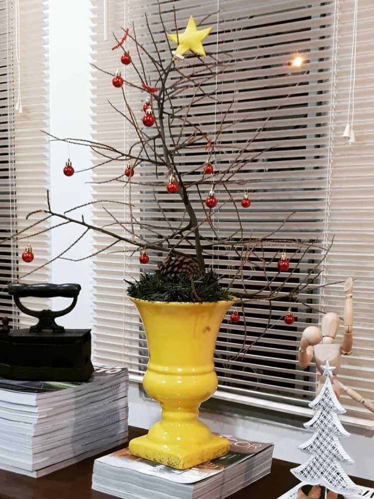 Árvore de Natal de galhos secos em vaso amarelo. Ela está cheia de enfeites vermelhos. 