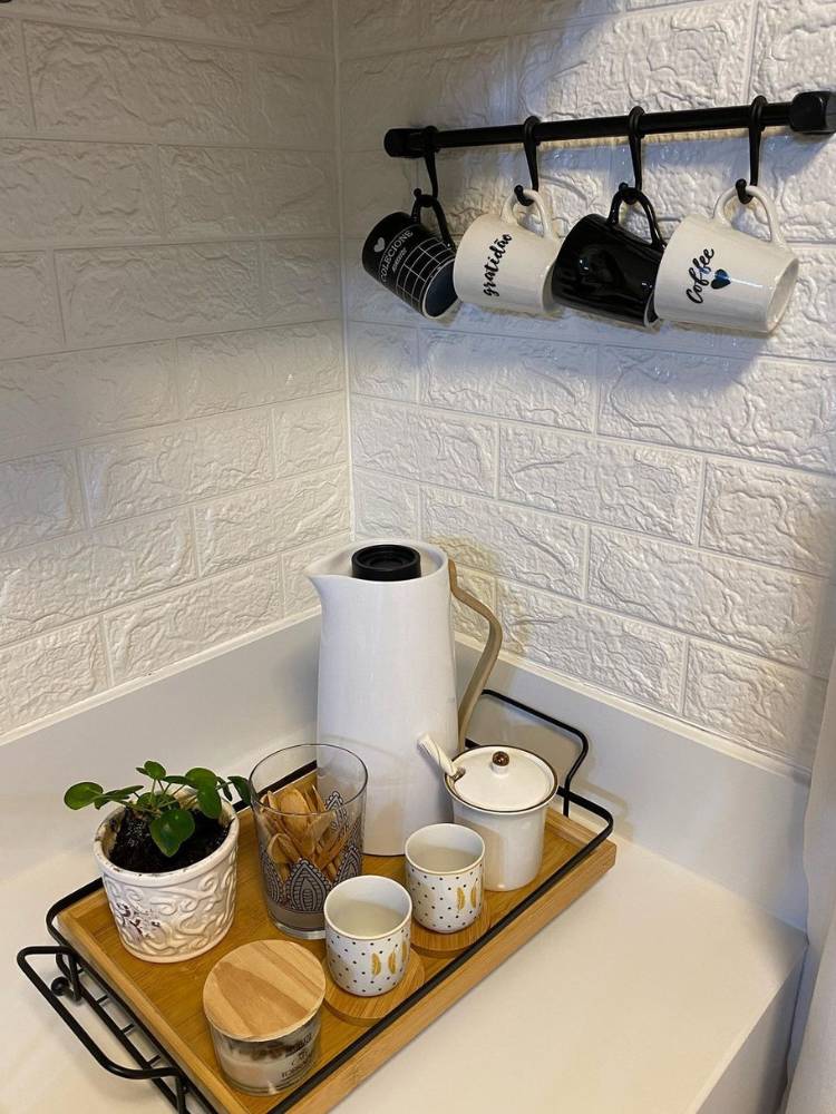 Cantinho do café em espaço de tijolinho à vista branco, com xícaras penduradas, bandeja marrom, garrafa térmica, potinhos e planta