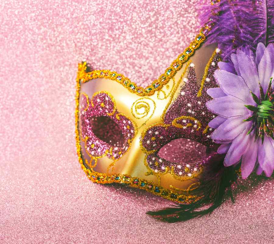 Foto de máscara de Carnaval dourada com detalhes em glitter e flor, sobre superfície pink brilhosa.