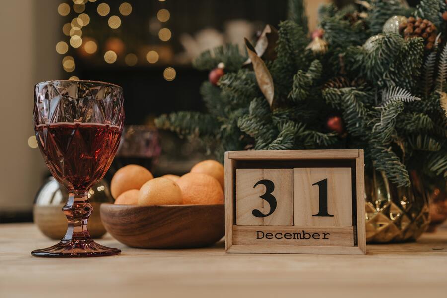 mesa de madeira com um calendário, também de madeira, mostrando que é dia 31 de dezembro. Do lado há laranjas, uma taça com bebida e, no fundo, uma árvore de natal