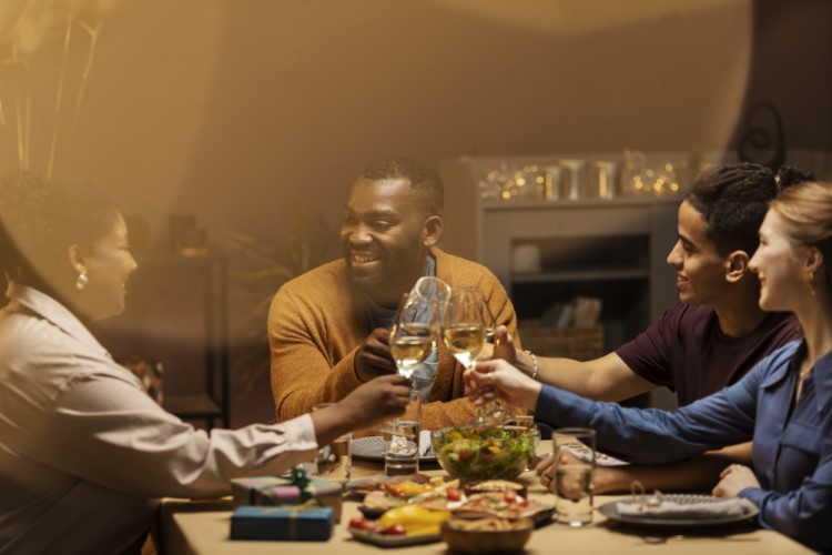 Quatro pessoas, sendo três de pele negra e uma de pele clara, sentadas à mesa repleta de alimentos e brindando. 