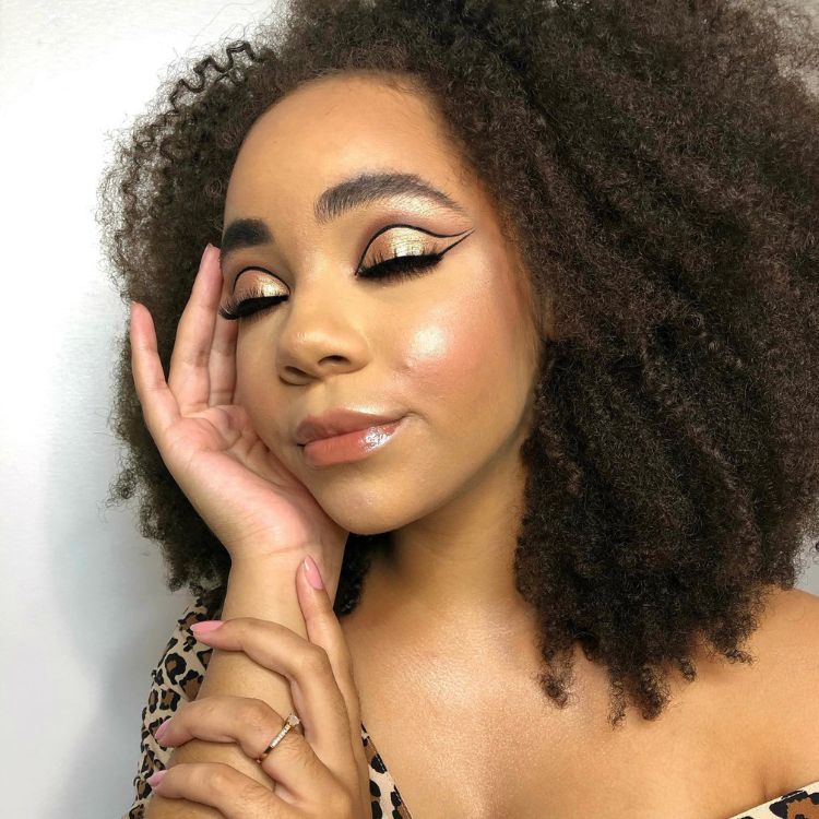 Mulher de pele negra usando maquiagem dourada com delineado gráfico preto, cílios postiço e gloss transparente.
