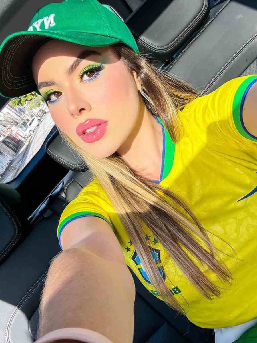 Selfie de mulher sentada em bando do carro usando camisa amarela do Brasil e boné verde.