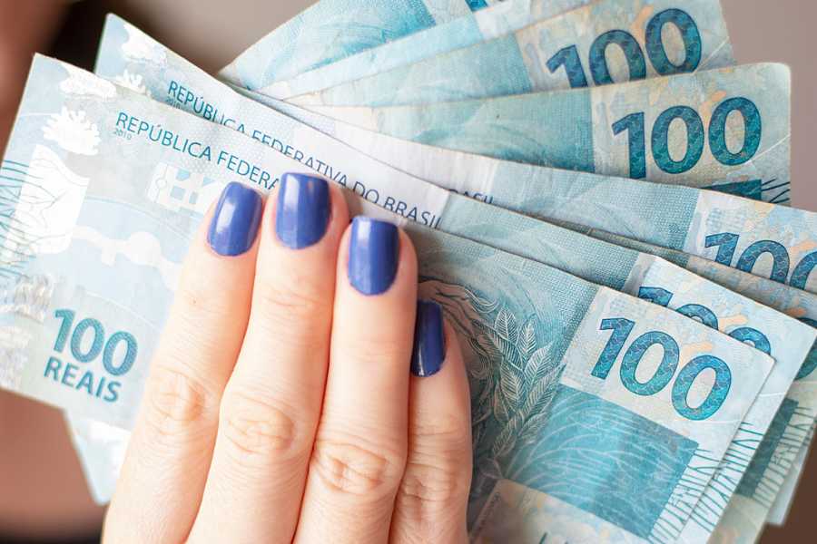 Mão de mulher segurando notas de R$ 100. 