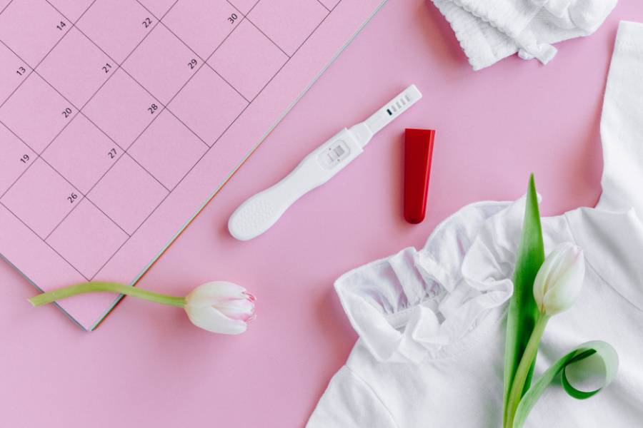 Teste de gravidez de farmácia em uma mesa rosa com flor e calendário ao redor.