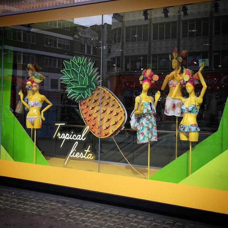 Vitrine de loja de moda praia chamada Tropical fiesta, com manequins de biquini e frutas na cabeça estilo Carmen Miranda. 