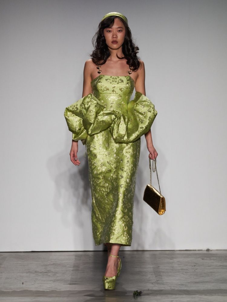 modelo com vestido midi verde, com babado na região do quadril, sapato e tiara também verdes e bolsa dourada.
