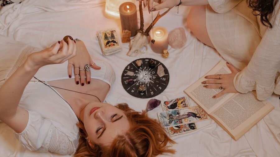 Garota ruiva deitada na cama, analisando um cristal com outra amiga do lado lendo tarot, com velas acesas e uma imagem similar a um horóscopo no meio dos lençóis brancos