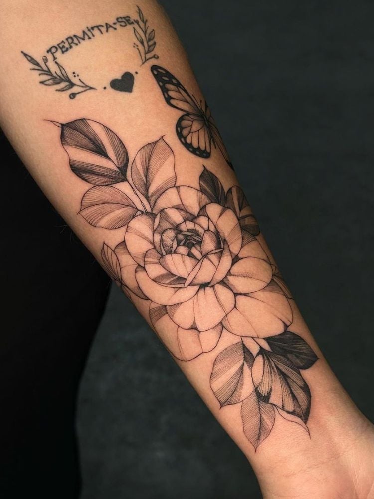 Foto de tatuagem no antebraço, com flor grande, borboleta e a frase "permita-se".  