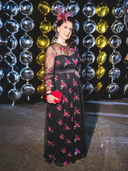 Na foto, Regina Caliman vestindo vestido longo preto, com bordados de flores. Adorno na cabeça e bolsa rosas. 