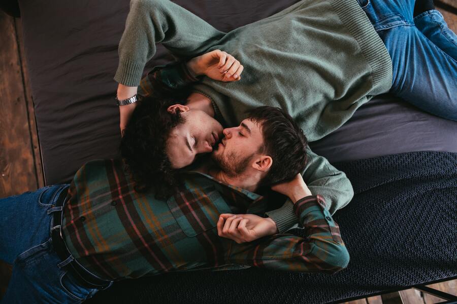 Casal branco homossexual com roupas escuras deitado em uma cama se beijando
