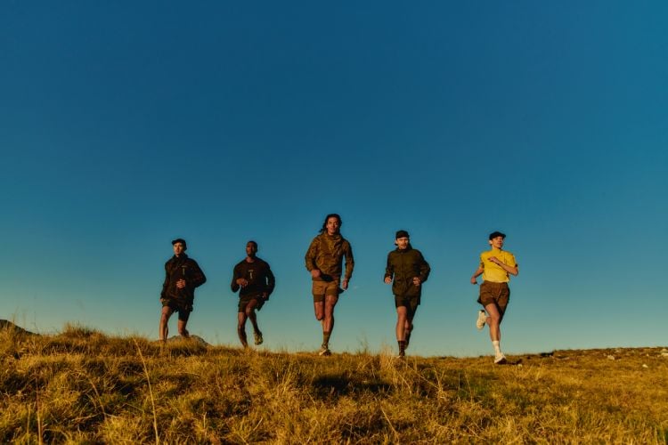 grupo de cinco pessoas com roupa de esporte e o novo tênis correndo em paisagem azul, chão de terra