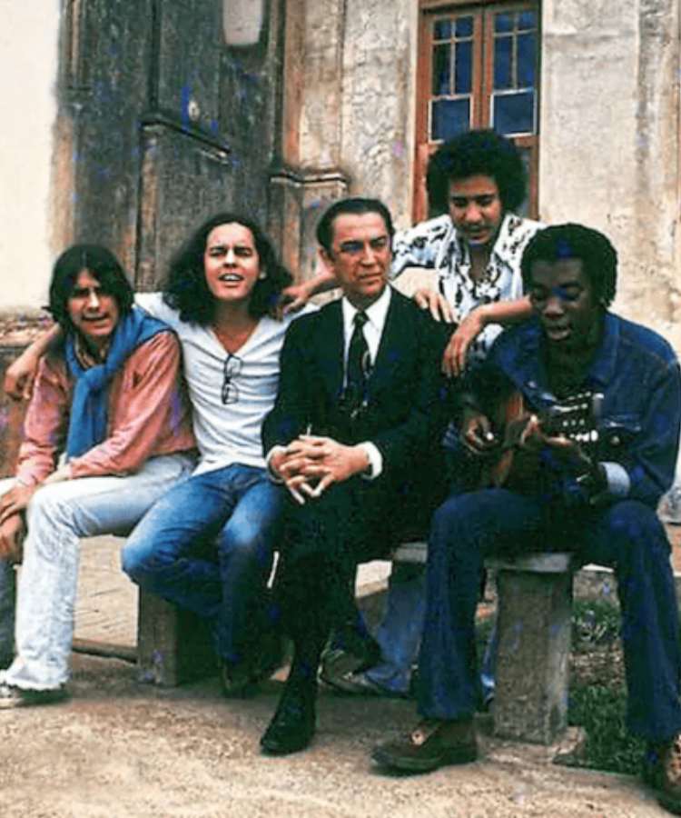 Integrantes do Clube de Esquina com Milton Nascimento tocando violão sentados em banco de rua.