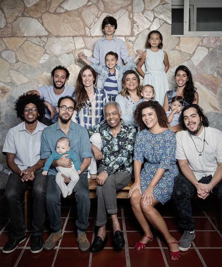 Cantor Gilberto Gil, seus filhos e filhas e netos e netas, sentados juntos de frente a parede de pedras.