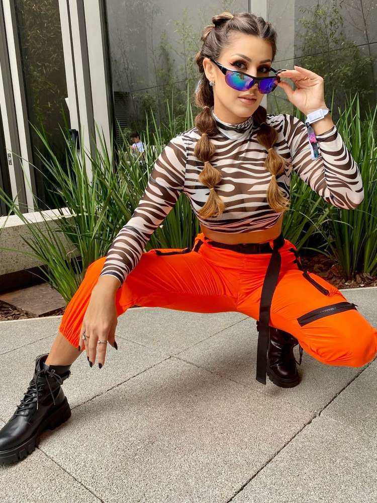 Mulher de pele clara usando cropped de zebra, óculos de sol, calça laranja e bota preta