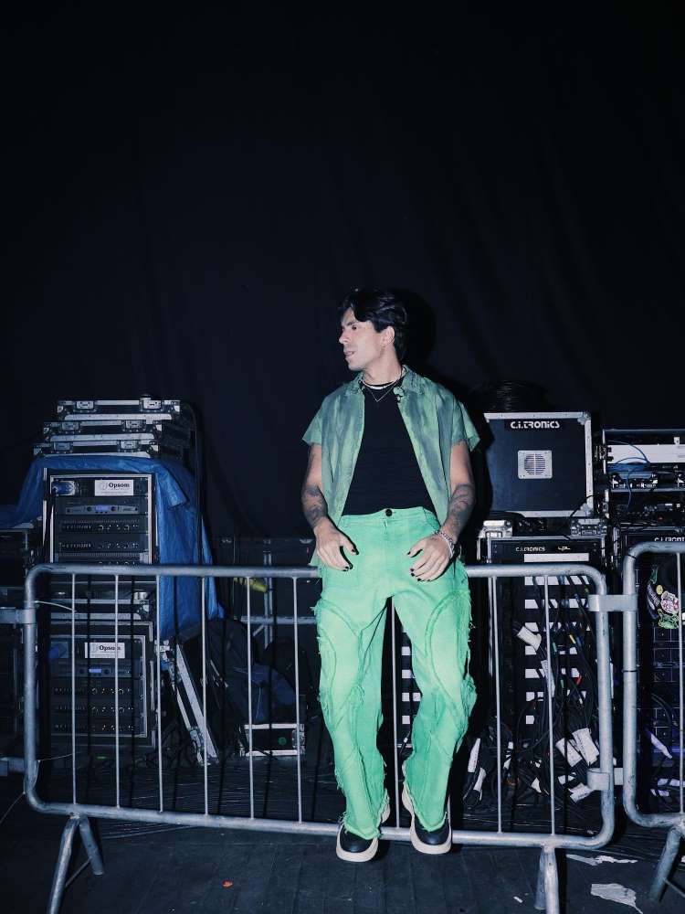 Pelu usando calça verde, blusa preta e camisa verde próximo à estrutura de palco