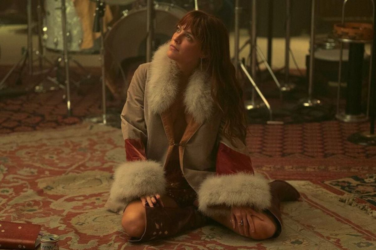 foto da personagem daisy jones sentada no chão, utilizando botas e casaco de pele.