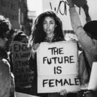 Mulher em protesto com outras mulheres, segurando placa escrito O futuro é feminoin em inglês