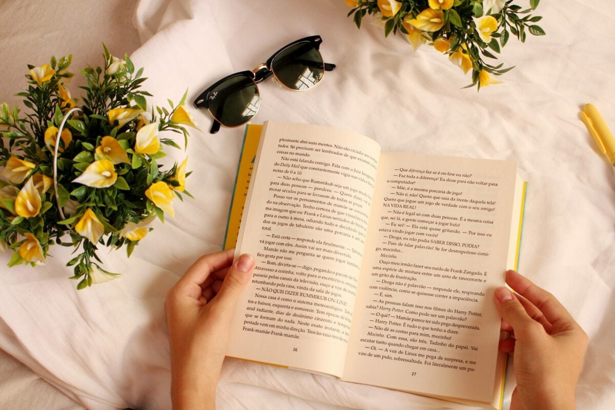 mão feminina segurando um livro com flores e um óculos do sol do lado