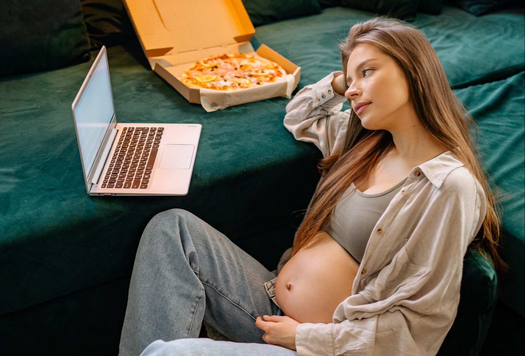 Mulher grávida lendo algo no notebook com uma caixa de pizza em cima do sofá