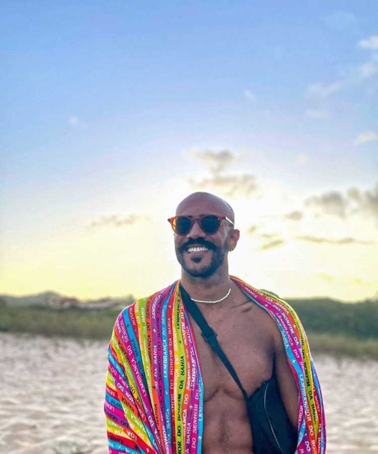Ricardo do BBB 23 de canga colorida nas costas e óculos de sol na praia.