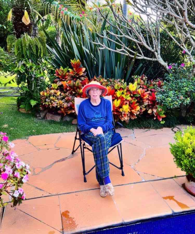 Cantora Rita Lee hoje em dia sentada em cadeira em local com flores e plantas coloridas.