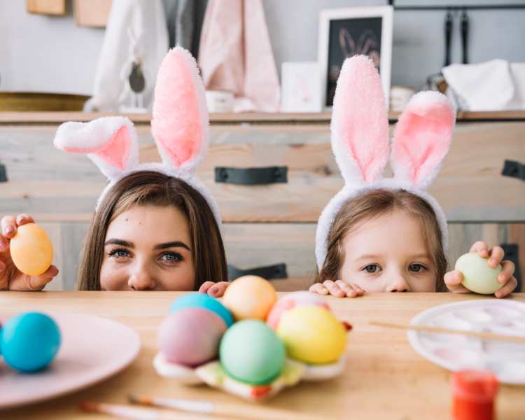 Menina e mãe com orelhas de coelho se escondendo atrás de mesa com ovos de galinha pintados para a Páscoa.