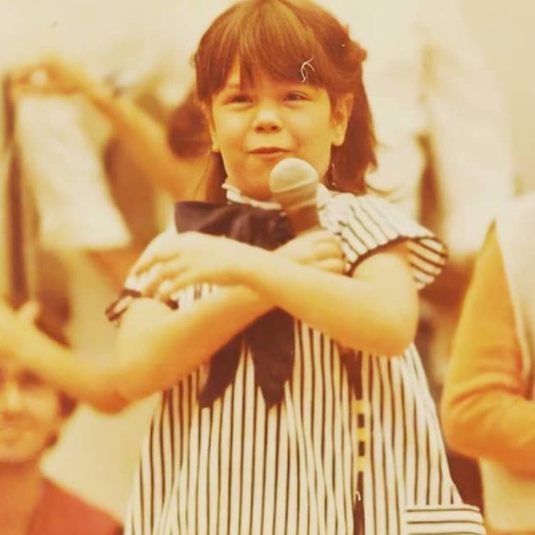 Uma foto nostálgica mostra a jovem segurando um microfone com as duas mãos, provavelmente durante sua época no grupo infantil Balão Mágico.