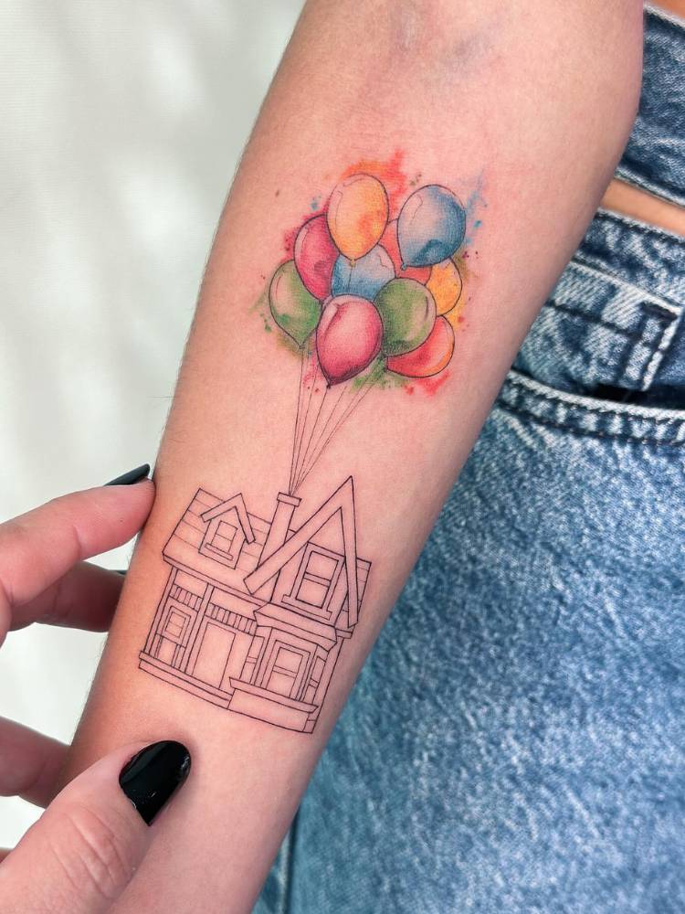 Tatuagem feminina no braço de Up Altas Aventuras, com uma casa em traços finos e balões coloridos. 