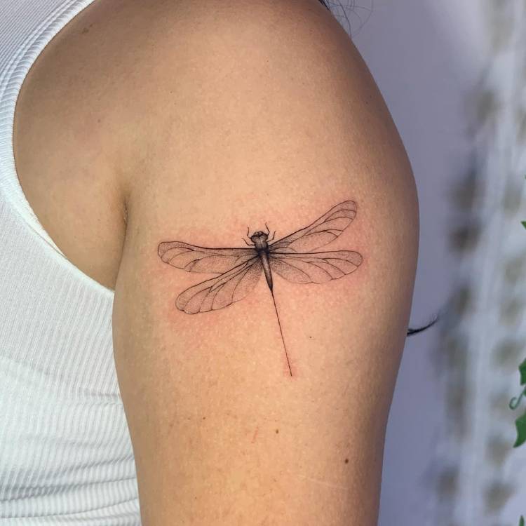 Tattoo de libélula no braço, próxima ao ombro, de uma mulher usando regata branca. 