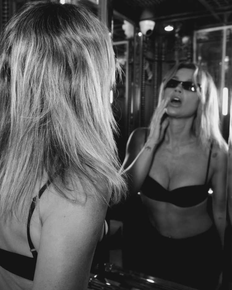 Foto em preto e branco de Flávia Alessandra com top preto e óculos escuro se olhando no espelho com mão perto da boca