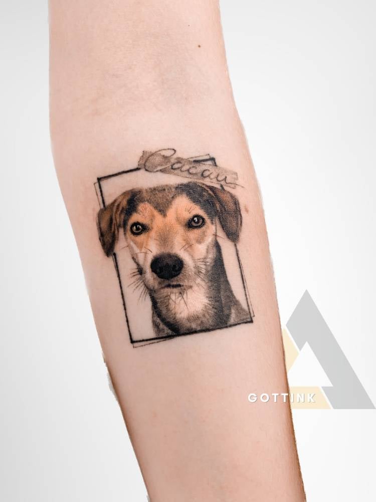 Tatuagem feminina realista de cachorro preto, branco e marrom, dentro de um retângulo e com o nome "Cacau". 