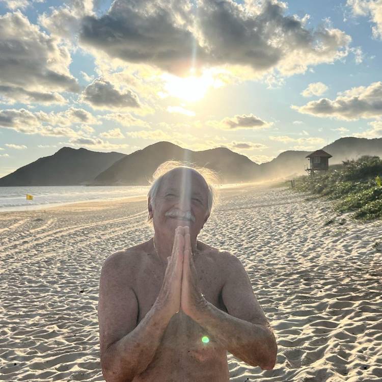 Ary Fontoura fazendo sinal de oração com as mãos em praia com vista em direção ao sol.