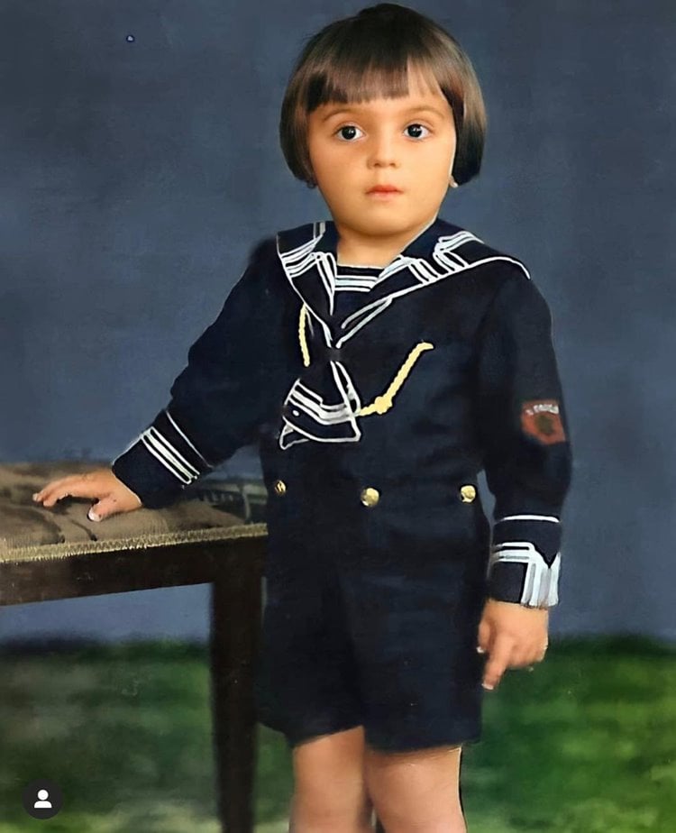 Ary Fontoura bebê, com roupa de marinheiro, encostando mãozinha em móvel com estofado.
