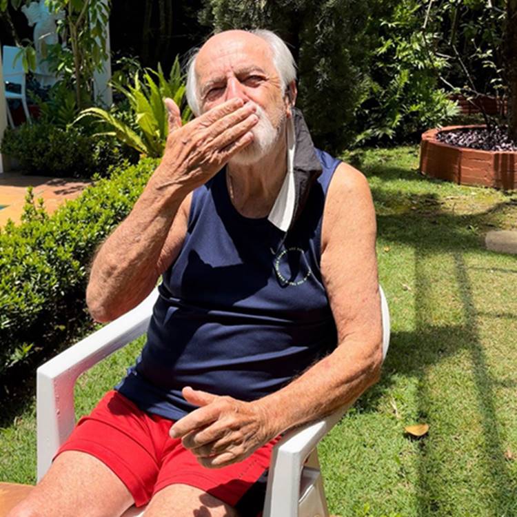 Ator Ary Fontoura sentado em cadeira de plástico em área externa de casa e mandando beijo com a mão. 