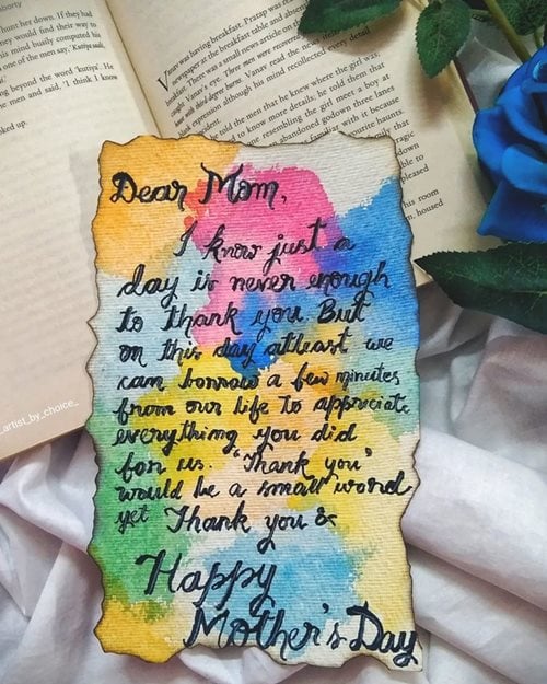 Carta feita a mão com texto em inglês para Dia das Mães, sobreposta sobre livro, tecido branco e rosa.