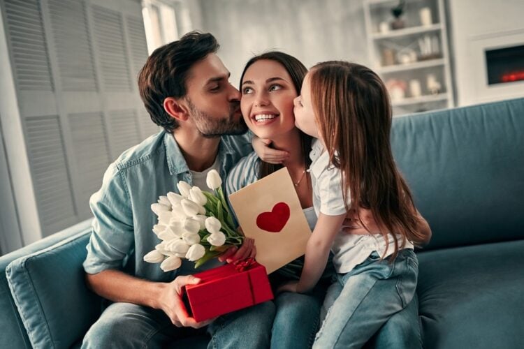 Foto de mãe recebendo beijo no rosto do marido e da filha. Ela segura presentes. Todos estão sentados em um sofá