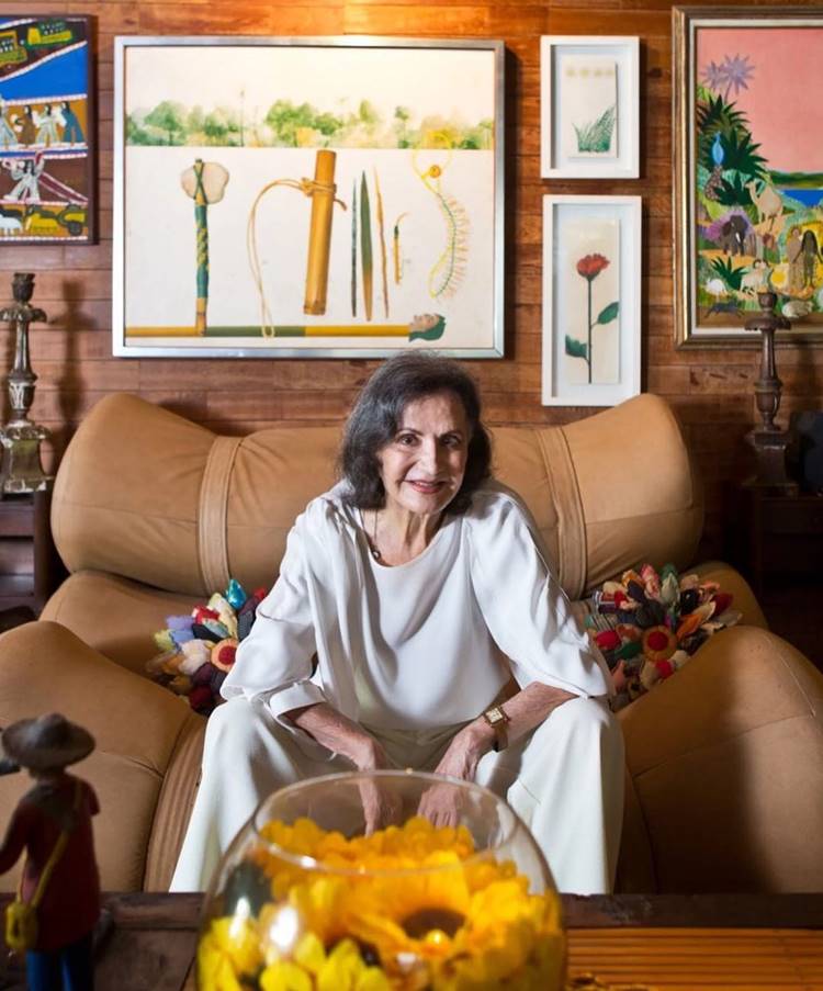 Rosamaria Murtinho hoje em dia, em foto sentada em estofado de casa com vários quadros na parede.