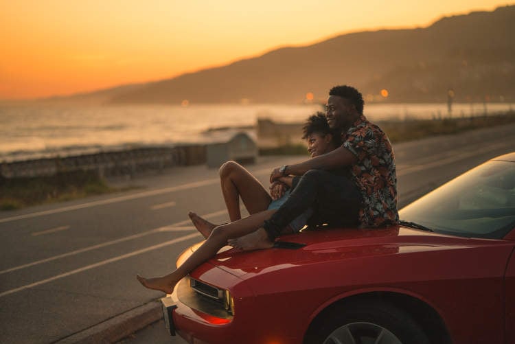 homem e mulher se abraçando em cima de um carro vermelho, enquanto olham o por d sol