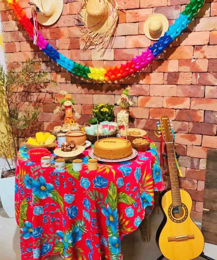Decoração em mesa de festa junina simples, contendo bolos, pipoca, milho, doces, violão, guirlanda, etc