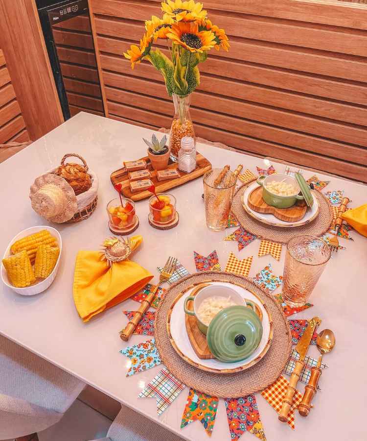 Decoração em mesa de festa junina, contém pratos, sousplat de bandeirinha, comidinhas como milho e salada de fruta,