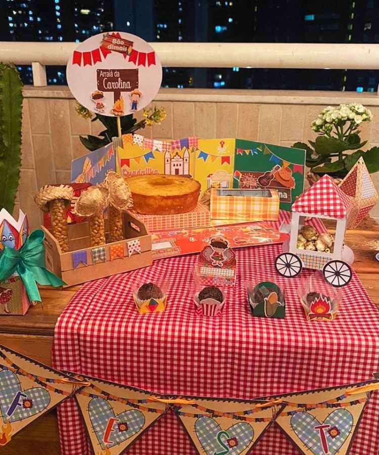 Decoração em mesa de festa junina simples, contendo doces, bolos, canecas, copos, enfeites de papel, etc